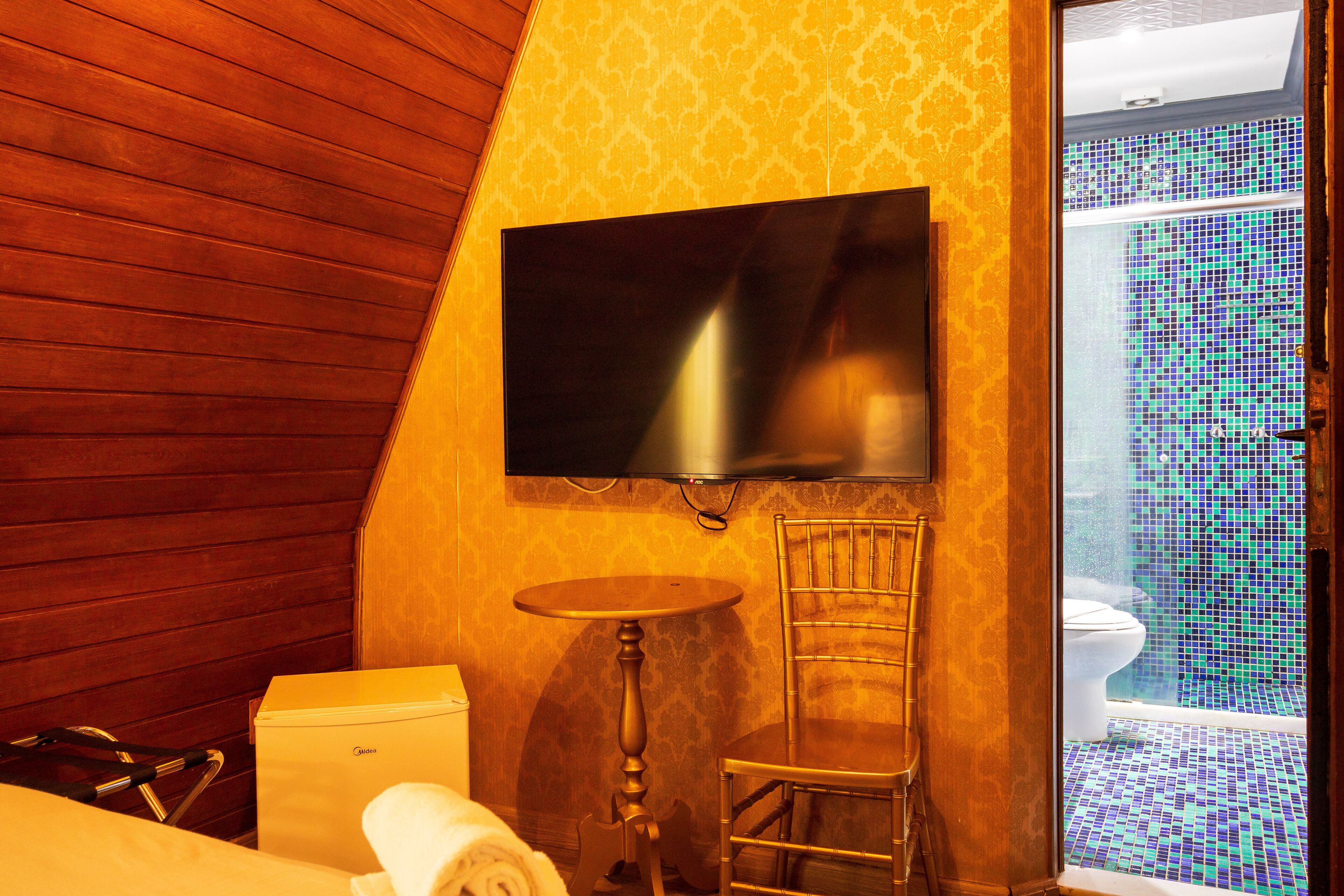 Ein Hotelzimmer mit schrägverkleidetem Holzboden, einem flachbildfernseher an der Wand, einem kleinen runden Tisch, einem Sessel, einem Bett mit einem aufgerollten weißen Handtuch und Blick auf ein Badezimmer mit Fliesenboden und Toilette.