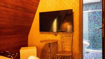 A hotel room with a wooden sloped ceiling, a flat-screen TV mounted on the wall, a small round table, a chair, a bed with a rolled-up white towel, and a view of a bathroom with a tiled floor and a toilet.