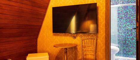 Ein Hotelzimmer mit schrägverkleidetem Holzboden, einem flachbildfernseher an der Wand, einem kleinen runden Tisch, einem Sessel, einem Bett mit einem aufgerollten weißen Handtuch und Blick auf ein Badezimmer mit Fliesenboden und Toilette.