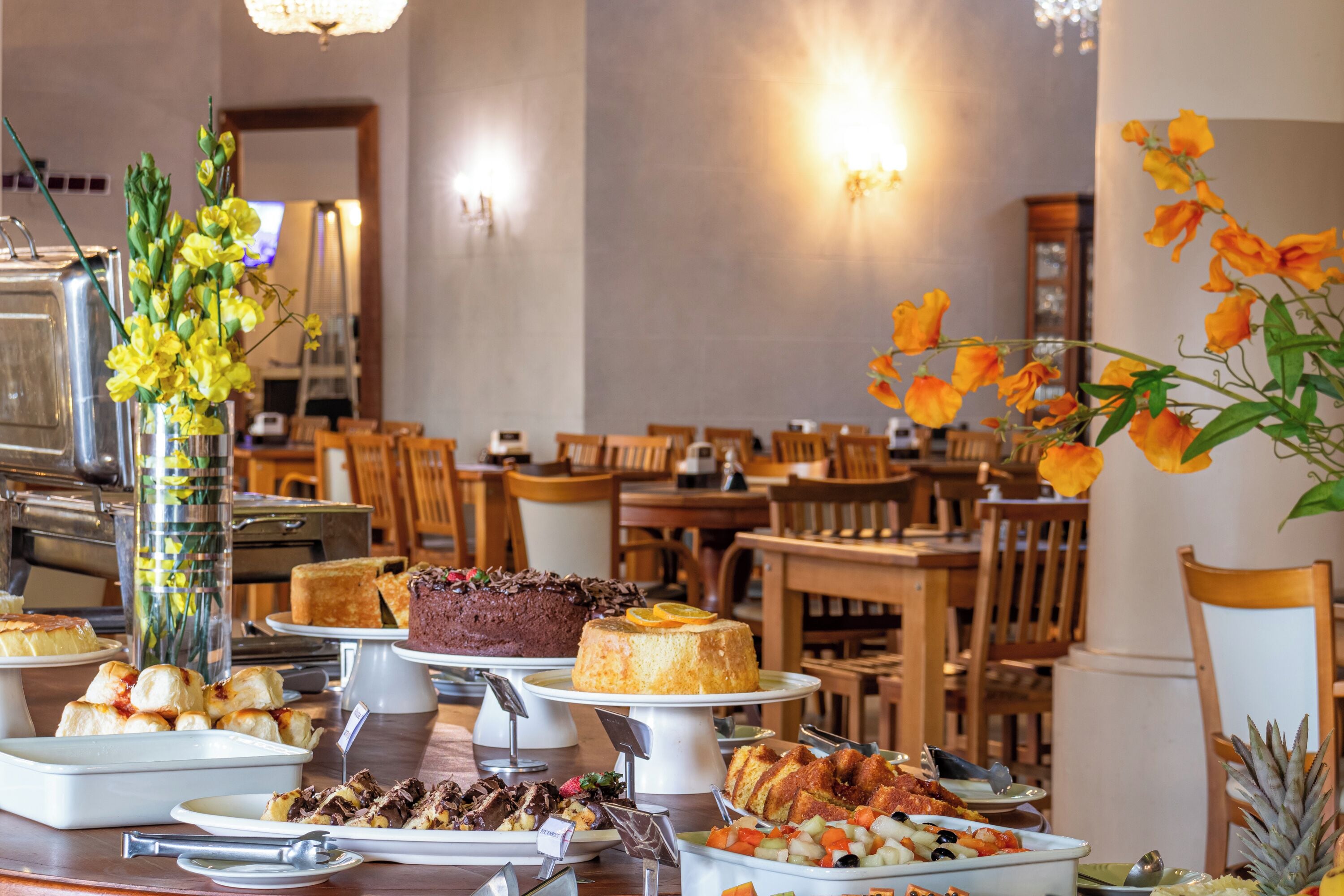 A buffet with various dishes, including cakes and fruits, in a dining area with wooden chairs and tables.