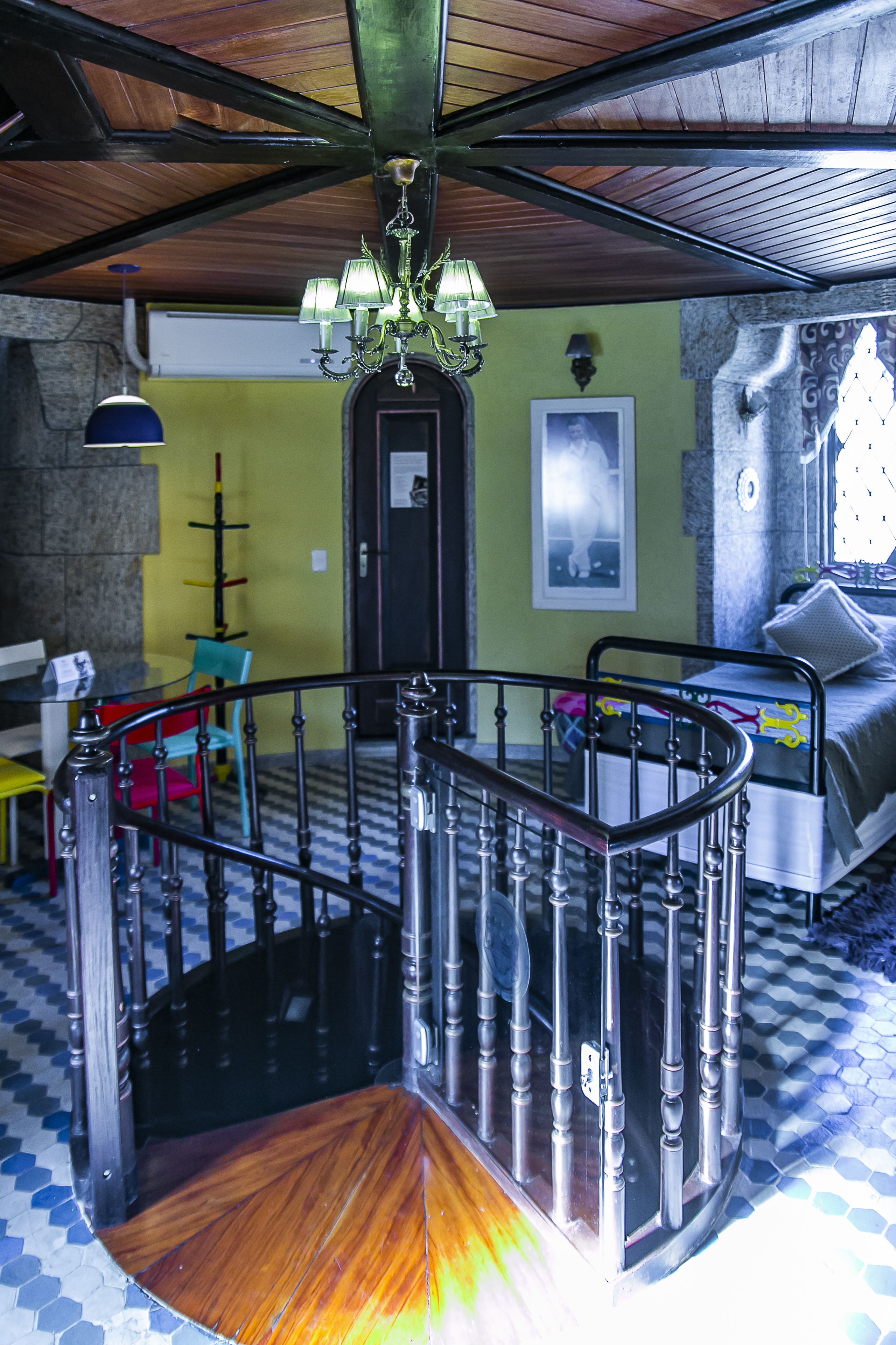 A spiral staircase with a wooden finish and metal balusters, leading to an upper floor. A chandelier hangs from the ceiling, and a bed with a patterned bedspread is visible on the upper floor.