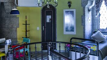 A spiral staircase with a wooden finish and metal balusters, leading to an upper floor. A chandelier hangs from the ceiling, and a bed with a patterned bedspread is visible on the upper floor.