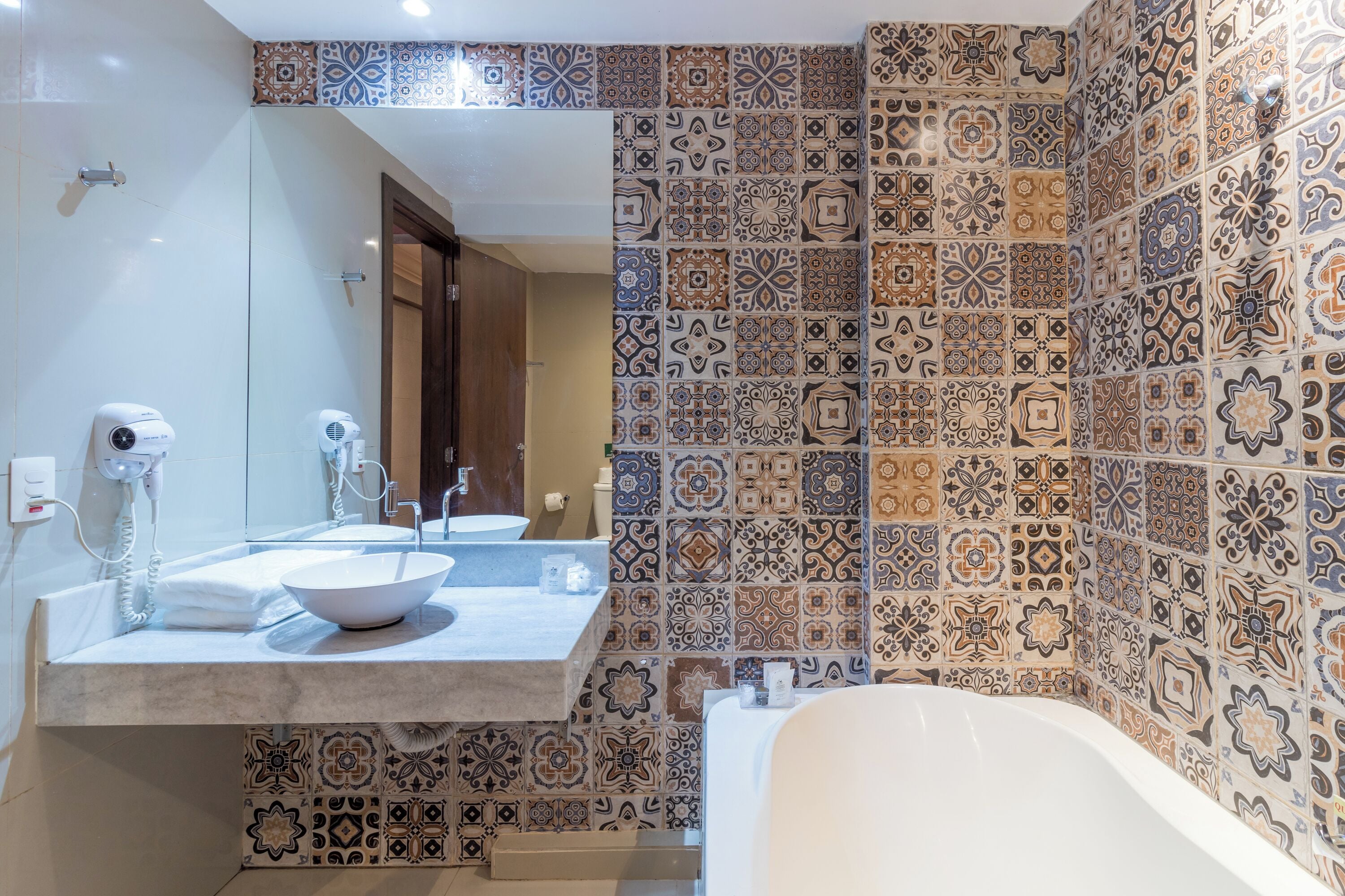 A bathroom with a large mirror, a sink with a bowl, a bathtub, and patterned tiles on the walls.