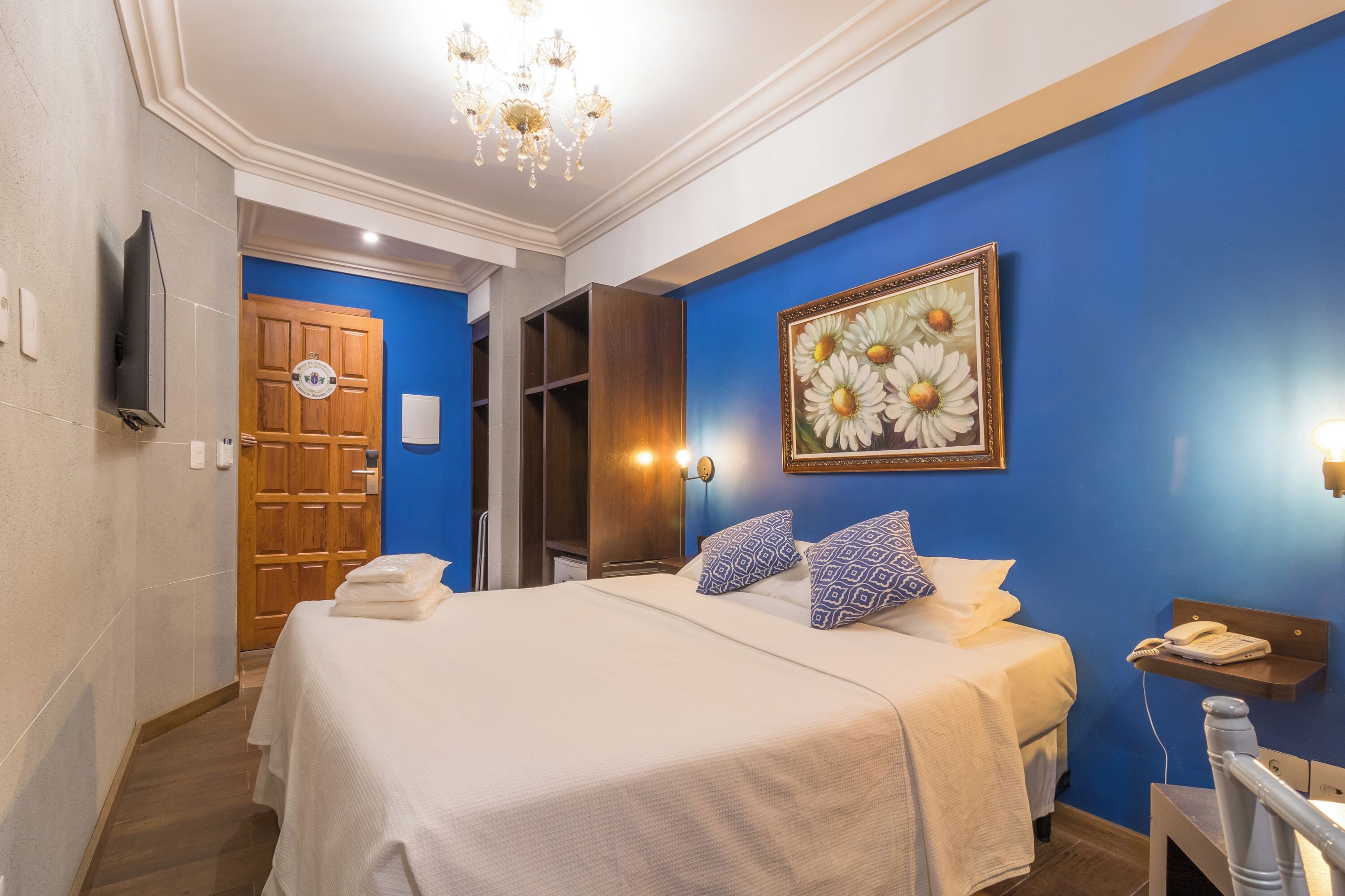 A hotel room with a large bed, a wooden wardrobe, a painting of daisies, and a chandelier.