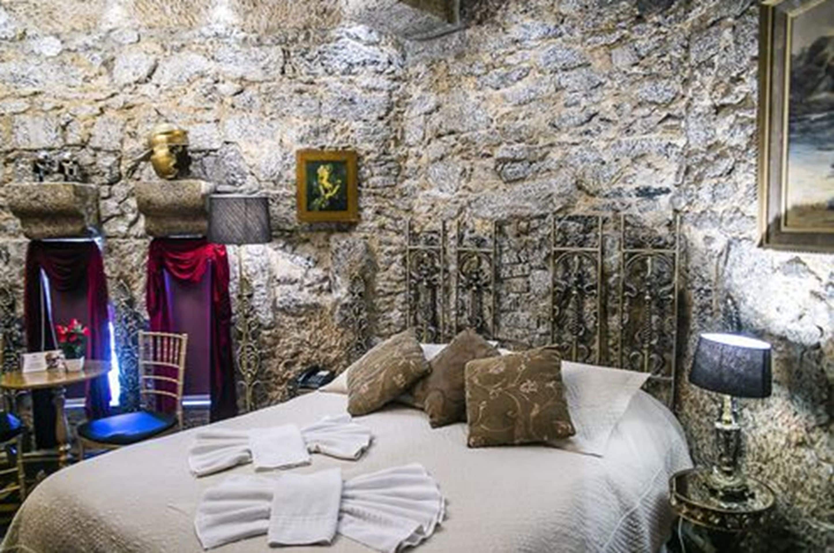 A stone-walled room with a bed, pillows, a lamp, and a framed picture on the wall.