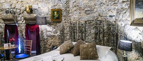 A stone-walled room with a bed, pillows, a lamp, and a framed picture on the wall.