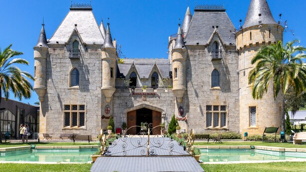 Un bâtiment qui ressemble à un château, avec une grande entrée centrale, de nombreuses tours et une piscine à l’avant.