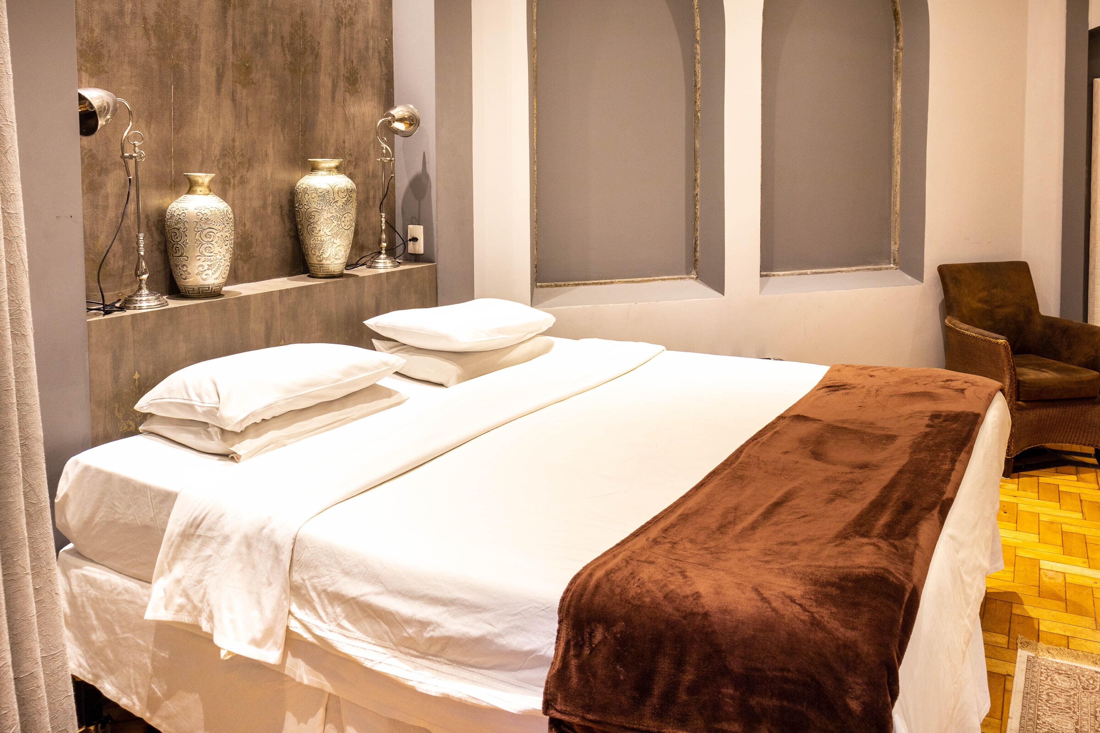 A neatly made bed with white linens and a brown throw blanket, a brown chair, and decorative vases on a shelf.