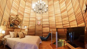 A room with a wooden sloped ceiling, a chandelier, a bed with pillows, a television, and a mirror. - Castelo de Itaipava - Hotel, Eventos e Gastronomia (Petrópolis)