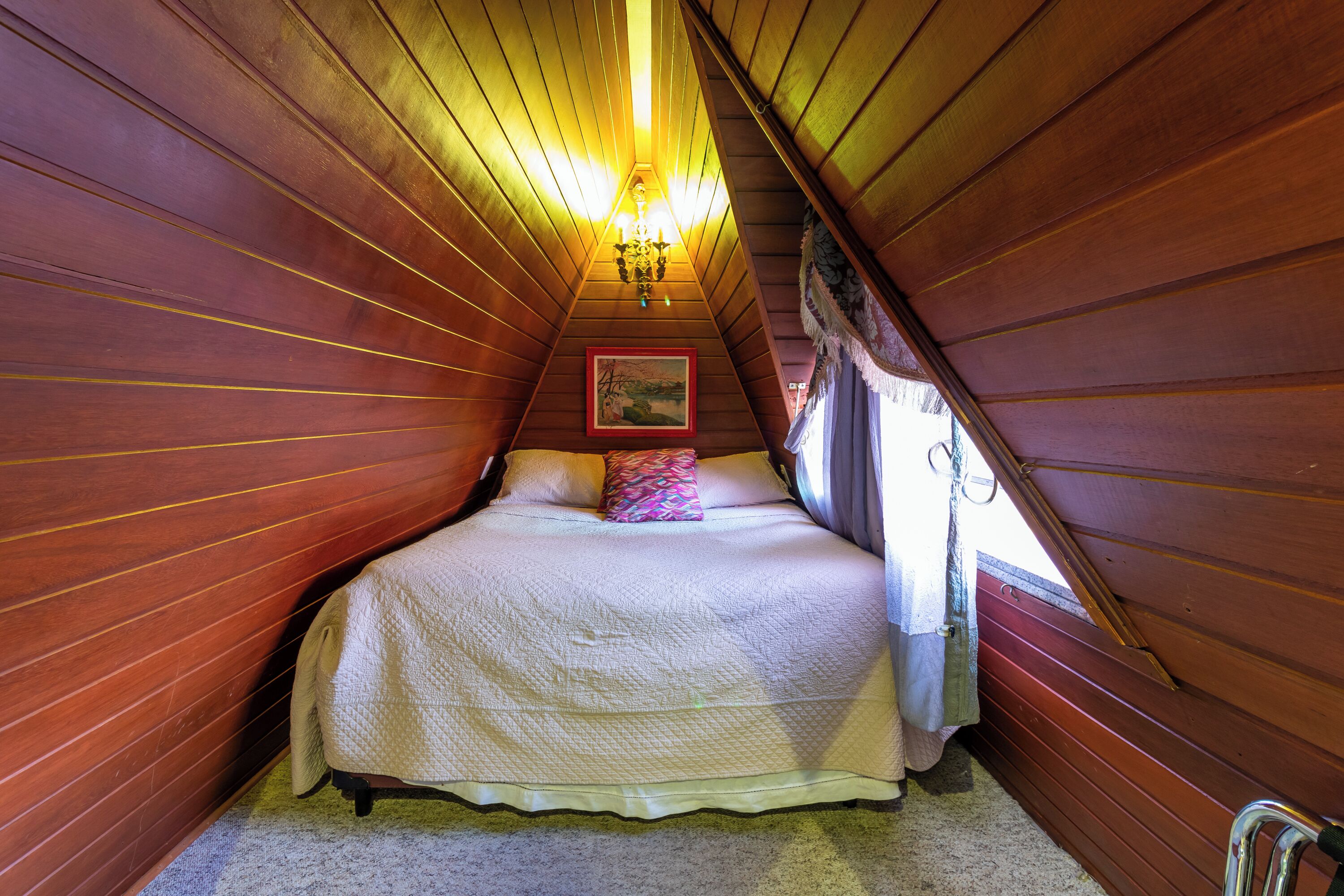 Un dormitorio con techo inclinado de madera, una cama con colcha blanca, un cojín colorido y un cuadro en la pared.