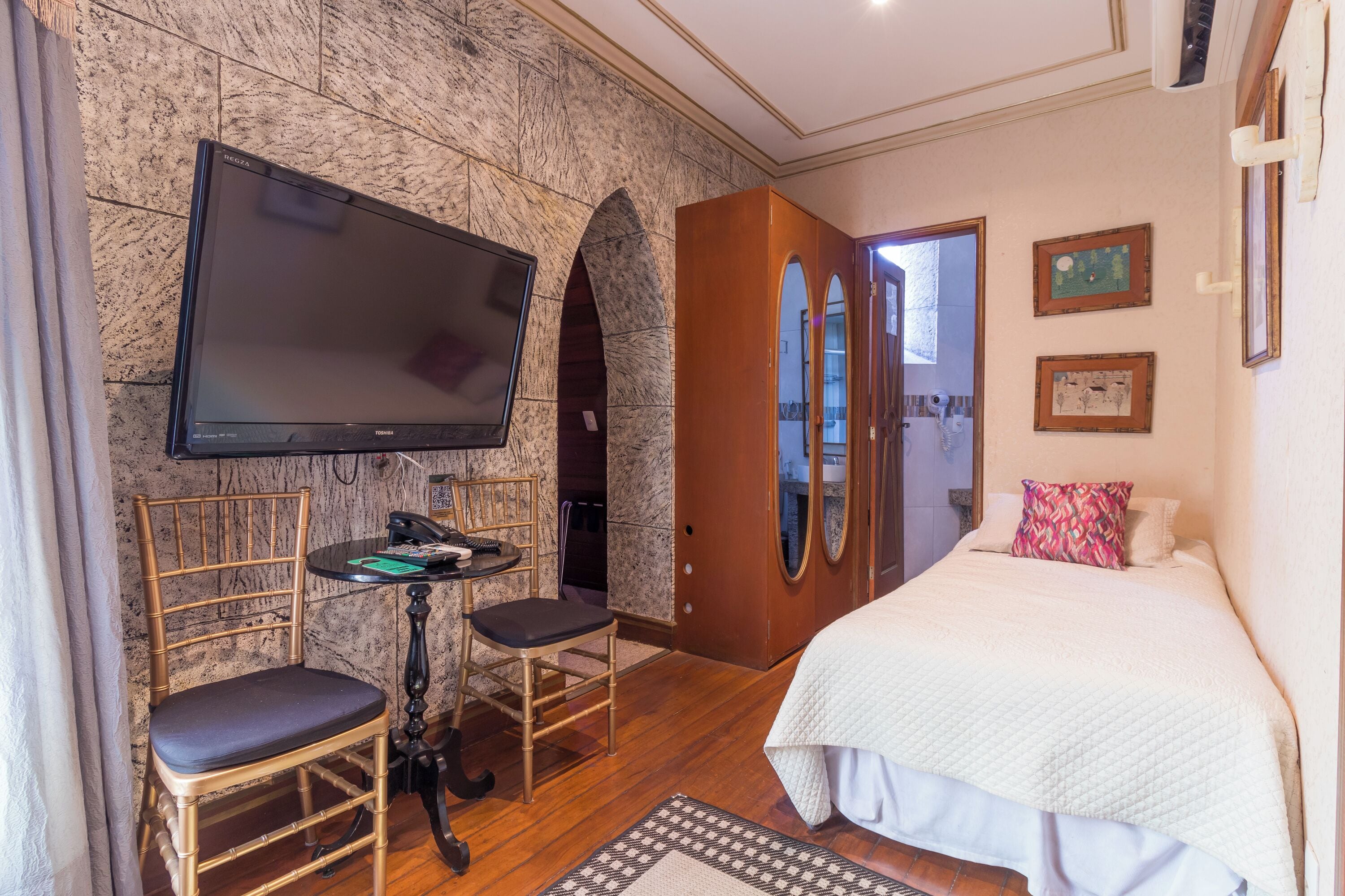 A hotel room with a bed, a TV mounted on a stone wall, a wooden chair, a small table, and a mirror.