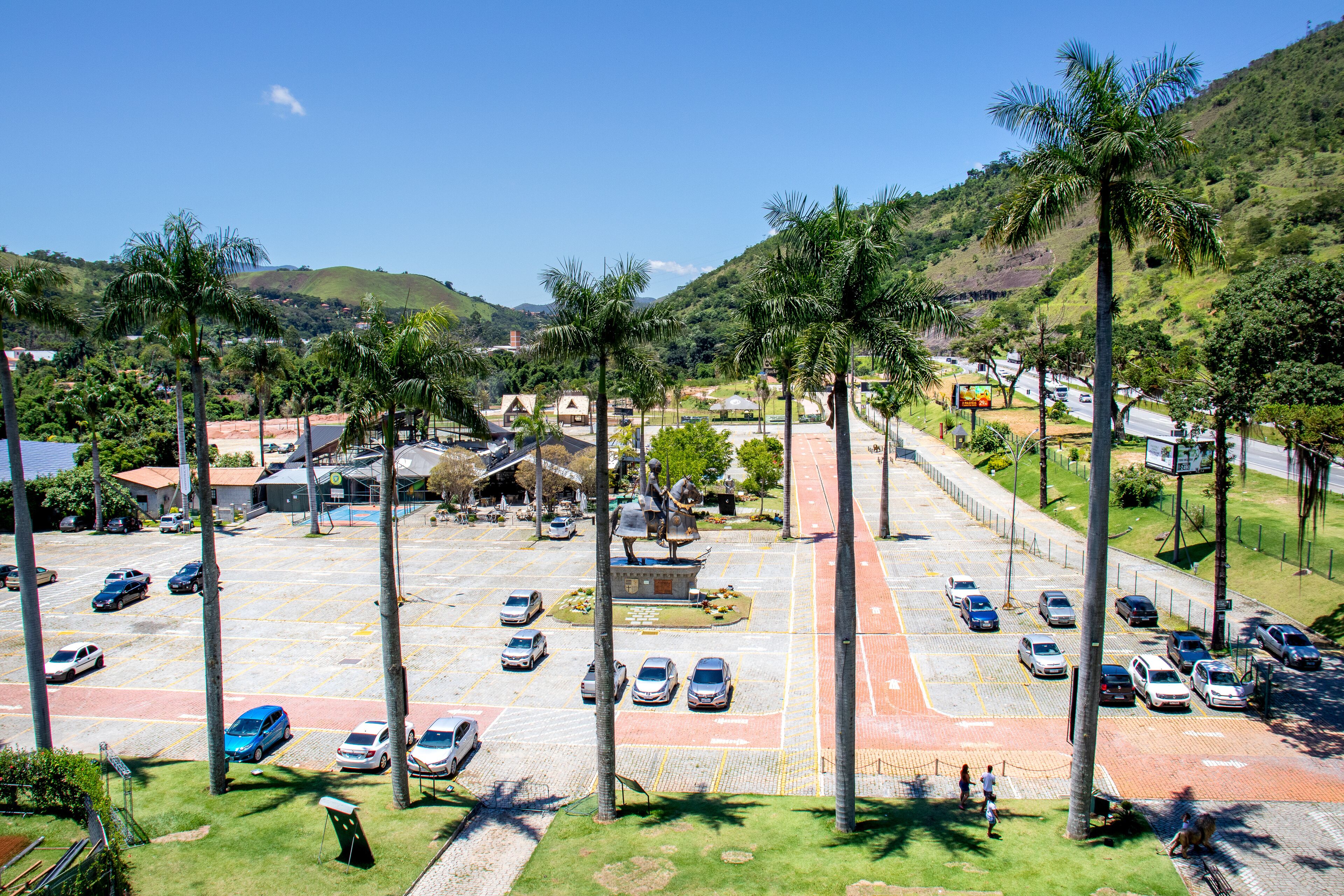 Un estacionamiento con varios coches, palmeras y un edificio con techo rojo al fondo.