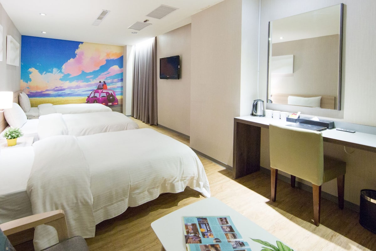 Quarto de hotel com duas camas, uma escrivaninha, um espelho, uma TV e um mural colorido na parede.
