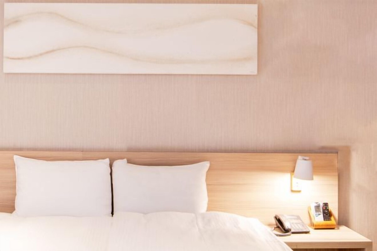 Una habitación de hotel con cabecera de madera, cama con ropa blanca, mesita de noche con lámpara y teléfono.