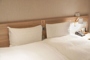 Habitación de hotel con cabecera de madera, dos almohadas blancas, una mesita de noche con lámpara, teléfono y reloj.