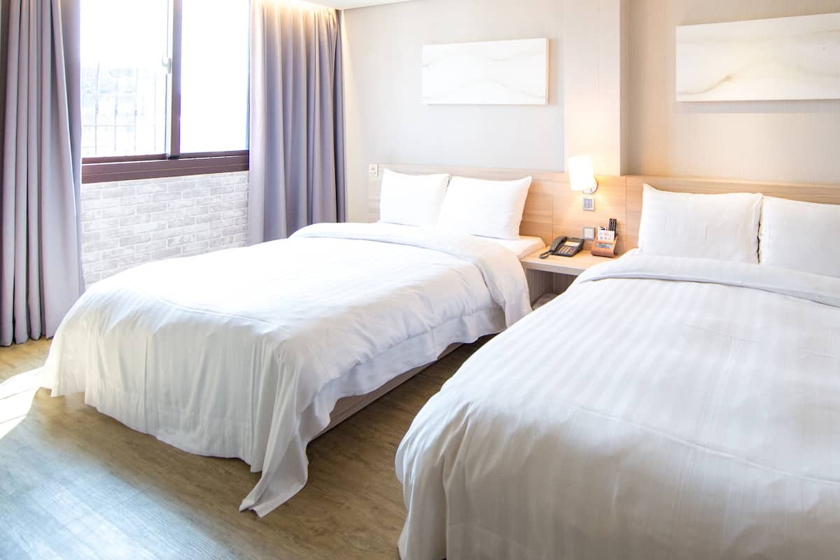 Quarto de hotel com duas camas, piso de madeira, parede de tijolos e janela com cortinas.