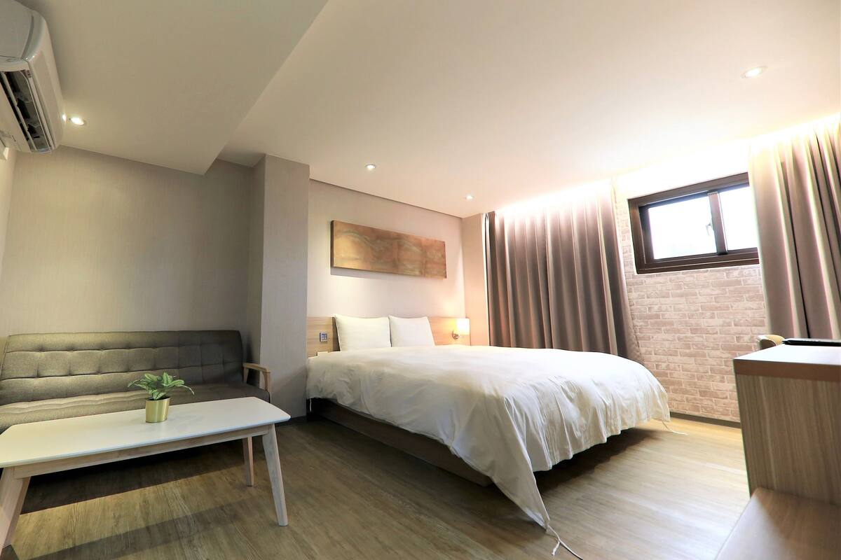 Quarto de hotel moderno com cama, escrivaninha pequena, sofá e planta em vaso.