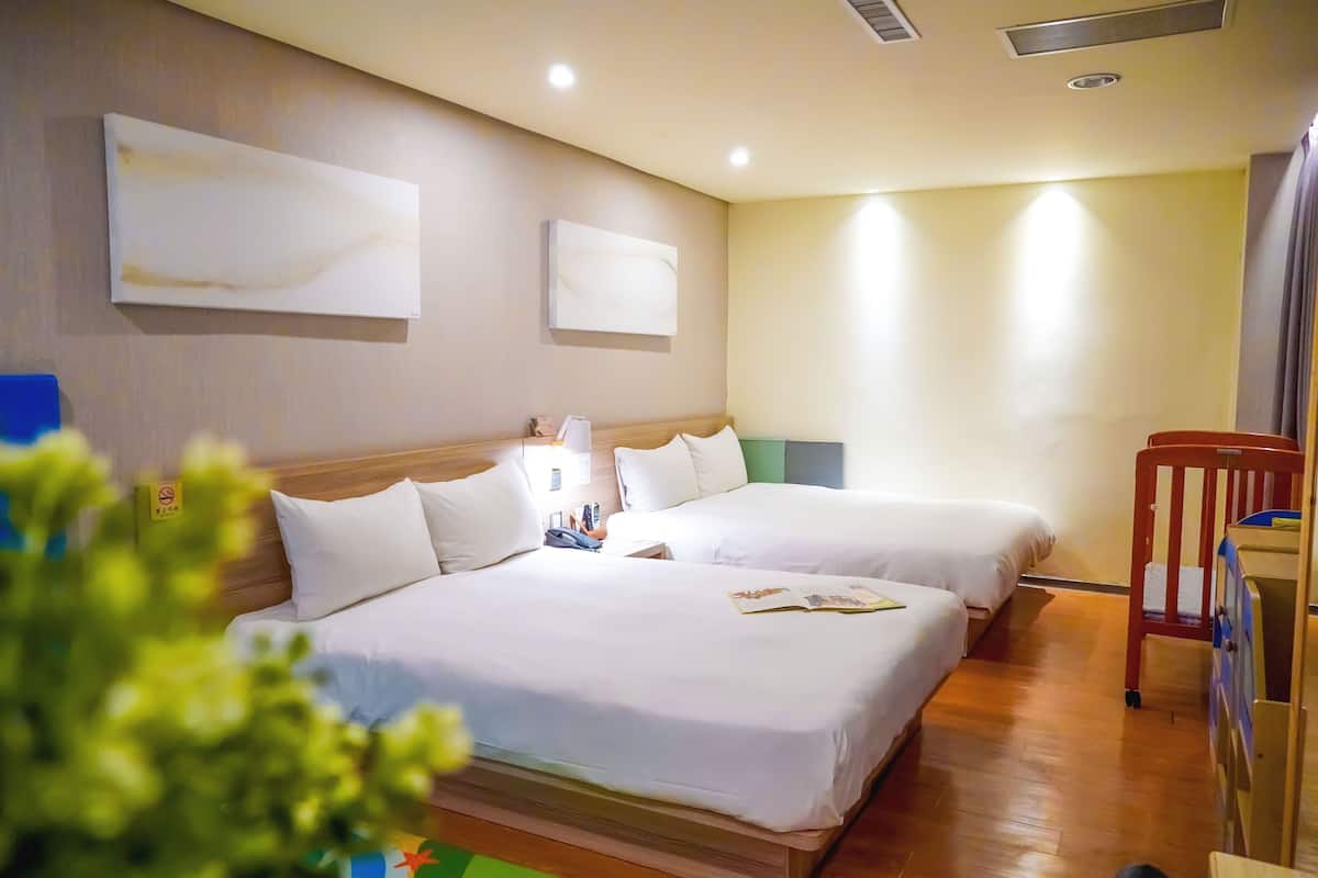 Quarto de hotel com duas camas, uma cabeceira de madeira, uma mesa de cabeceira com abajur e uma escrivaninha com cadeira.