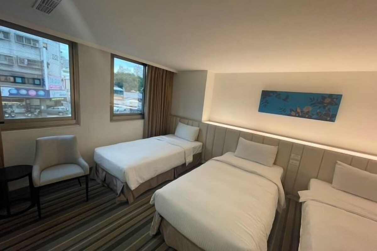 A hotel room with two single beds, a chair, a small table, and a large window offering a city view.