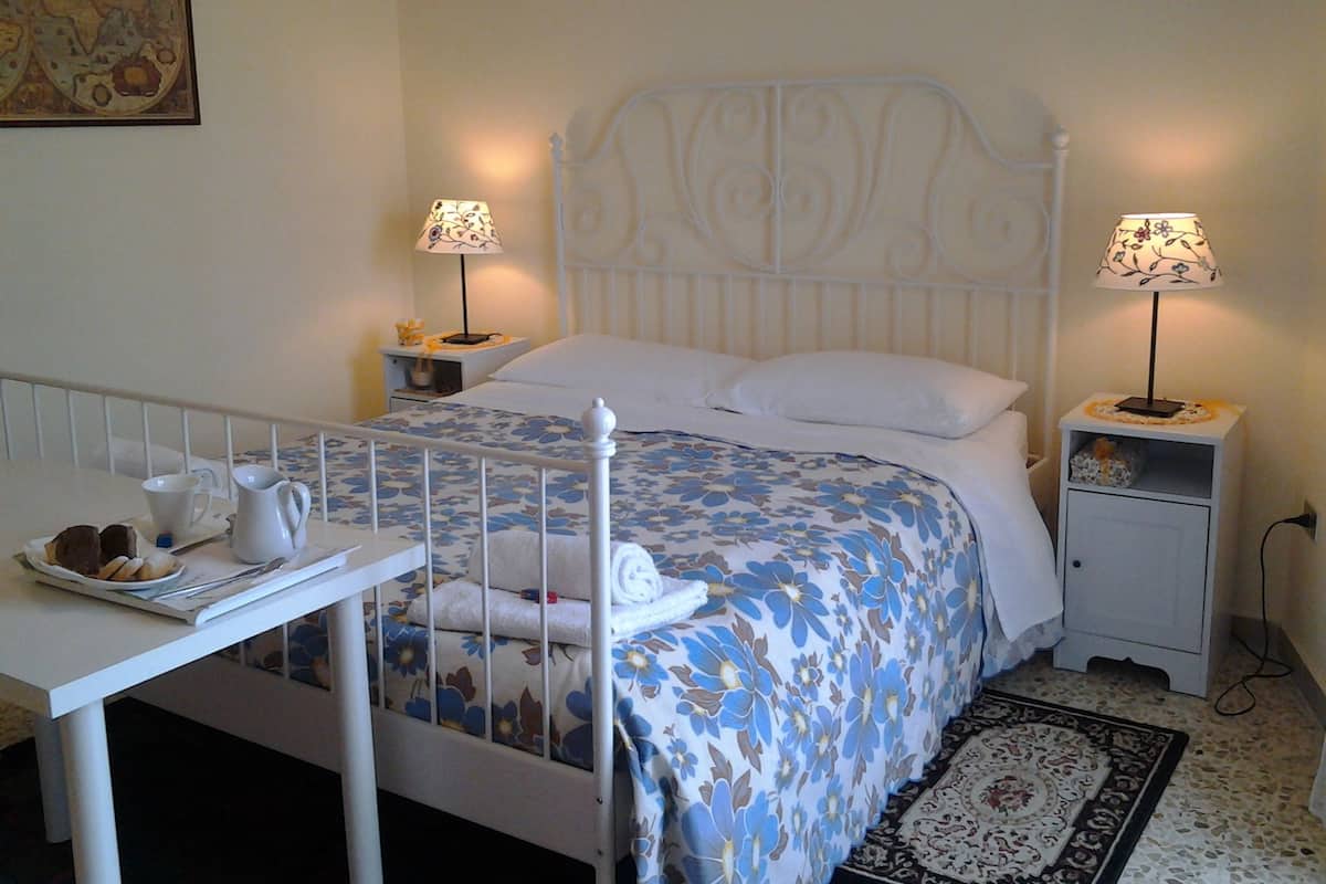Cama bem arrumada com colcha floral, duas mesas de cabeceira com abajures, uma mesinha com bandeja de doces e um tapete decorativo.