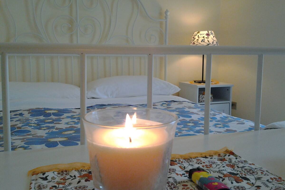 Quarto com cama, mesa de cabeceira com abajur e uma vela acesa sobre uma mesa.