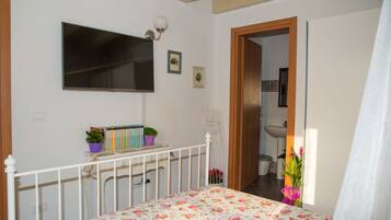 Un dormitorio con cama, televisión, mesita de noche con libros y una planta, y un baño visible a través de una puerta abierta.