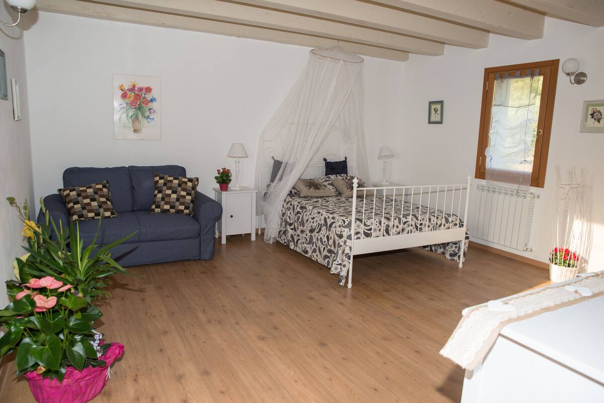 A bedroom with a bed, a sofa, a nightstand, a bedside table, a window, and a potted plant.