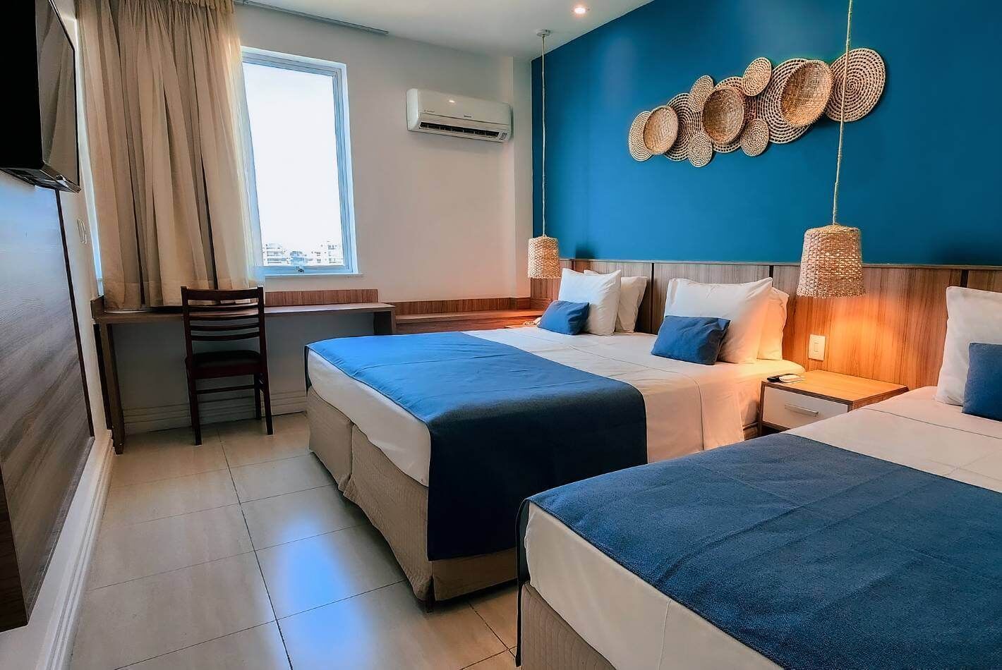 Quarto de hotel com duas camas, uma escrivaninha, uma cadeira e uma janela grande.