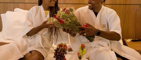 Une chambre d’hôtel avec un lit, un plateau de fruits et de desserts, et un bouquet de fleurs.