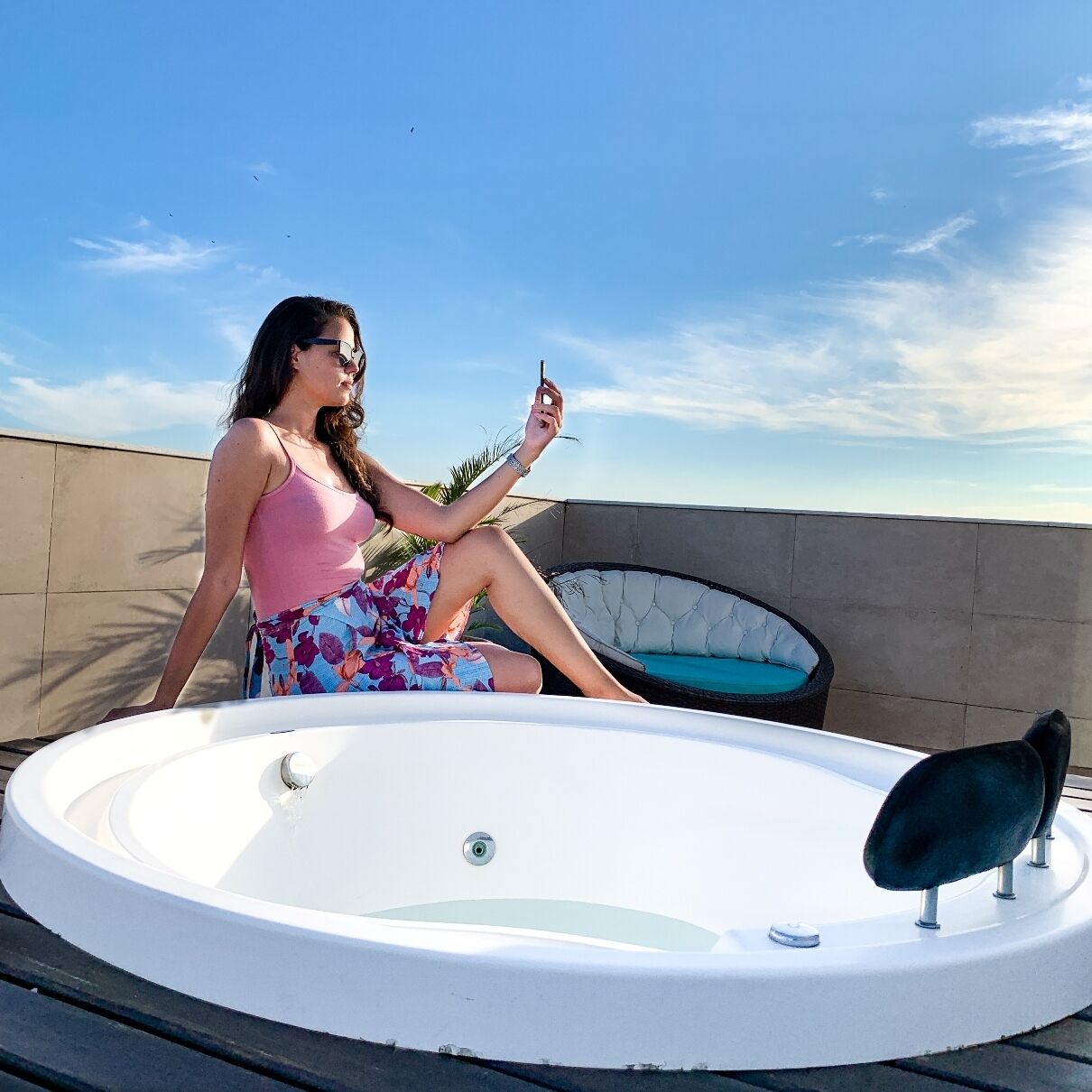 Uma pessoa sentada perto de uma banheira de hidromassagem circular em um terraço no telhado, com um céu claro ao fundo.