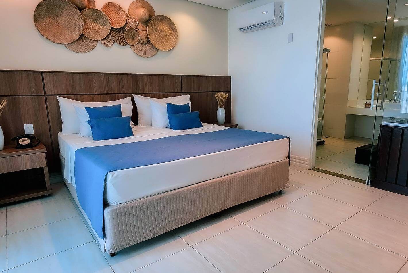 A modern hotel room with a large bed, a wooden headboard, a wall-mounted air conditioner, and a glass shower enclosure.