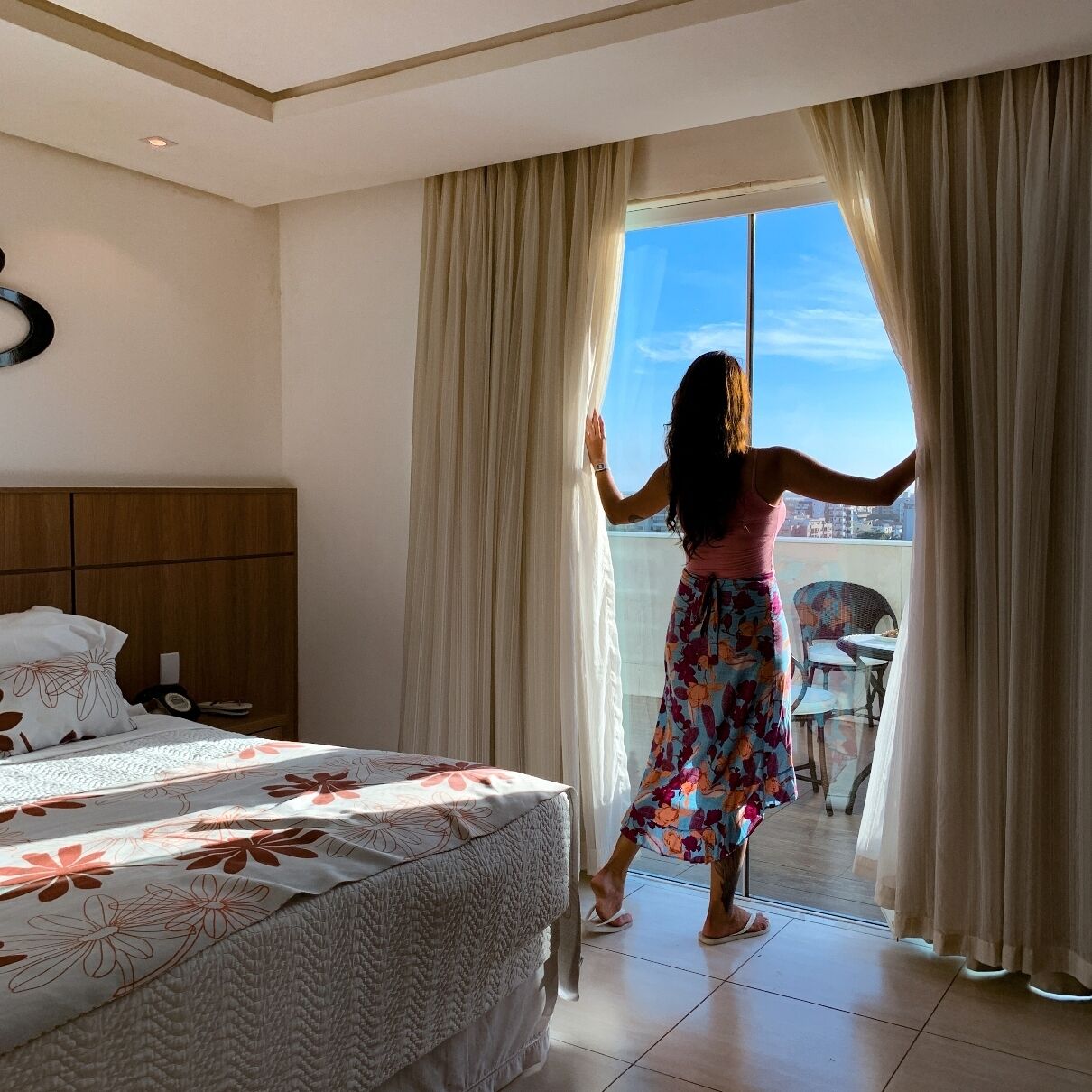 A hotel room with a bed, a wooden headboard, and a balcony with a view of the city.