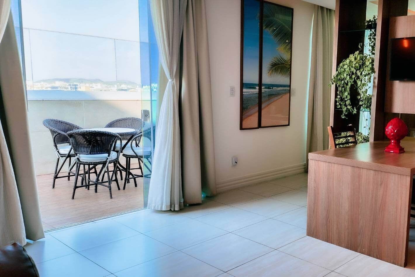 A room with a balcony, a dining area with wicker chairs, a wooden desk, and a framed picture of a beach.