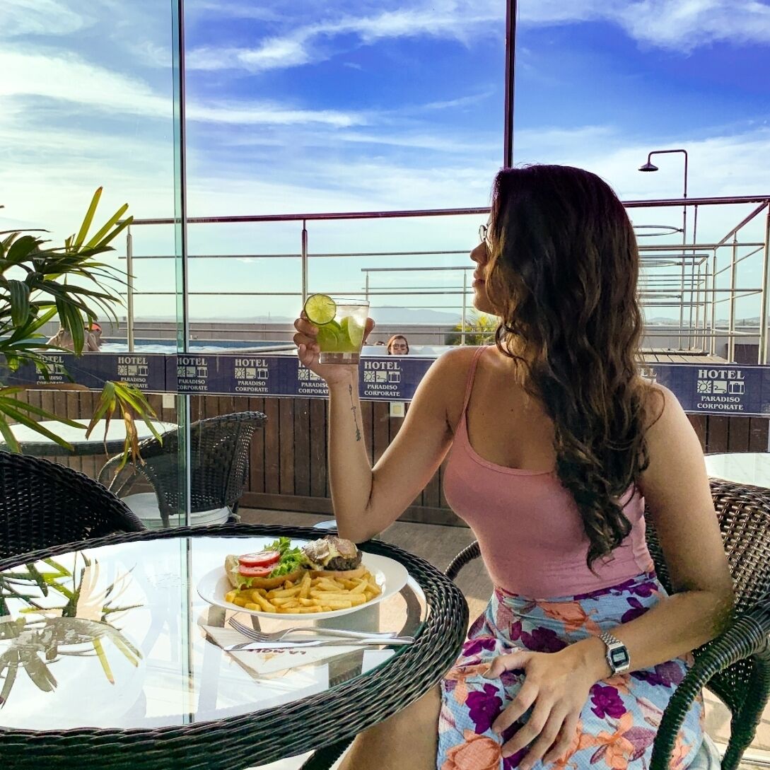 Una mujer sentada a una mesa con comida y bebida, con vistas a un paisaje con varios carteles que indican “HOTEL PARADISO CORPORATE”.