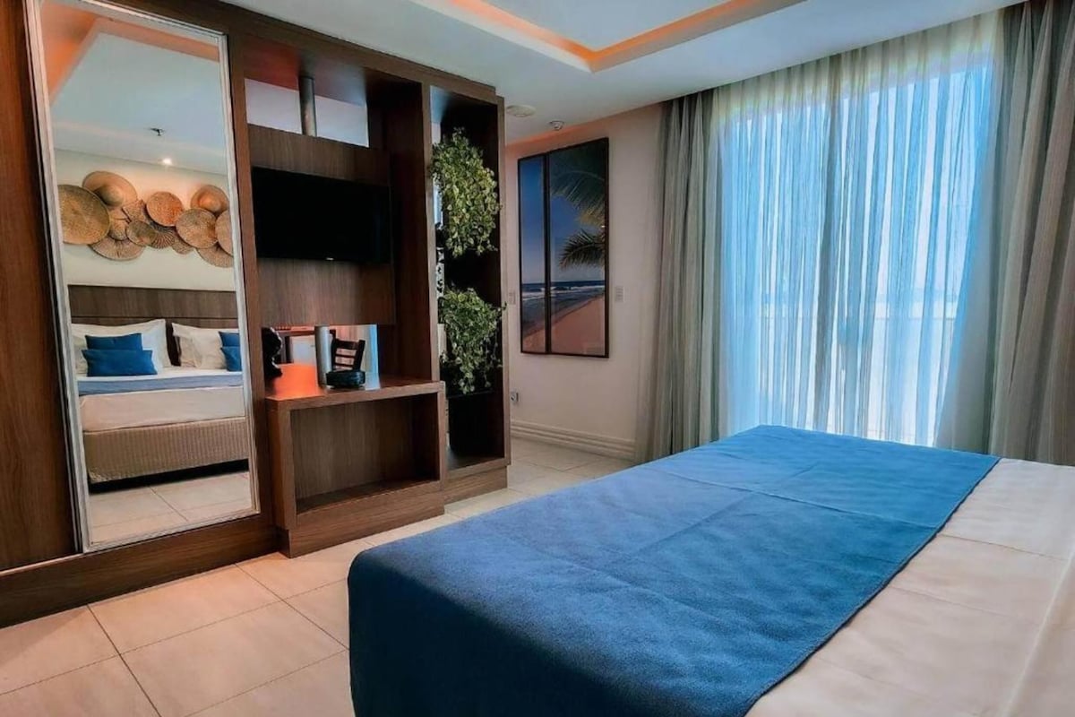 A hotel room with a bed, a wardrobe, a shelf with decorative items, and a large window with curtains.