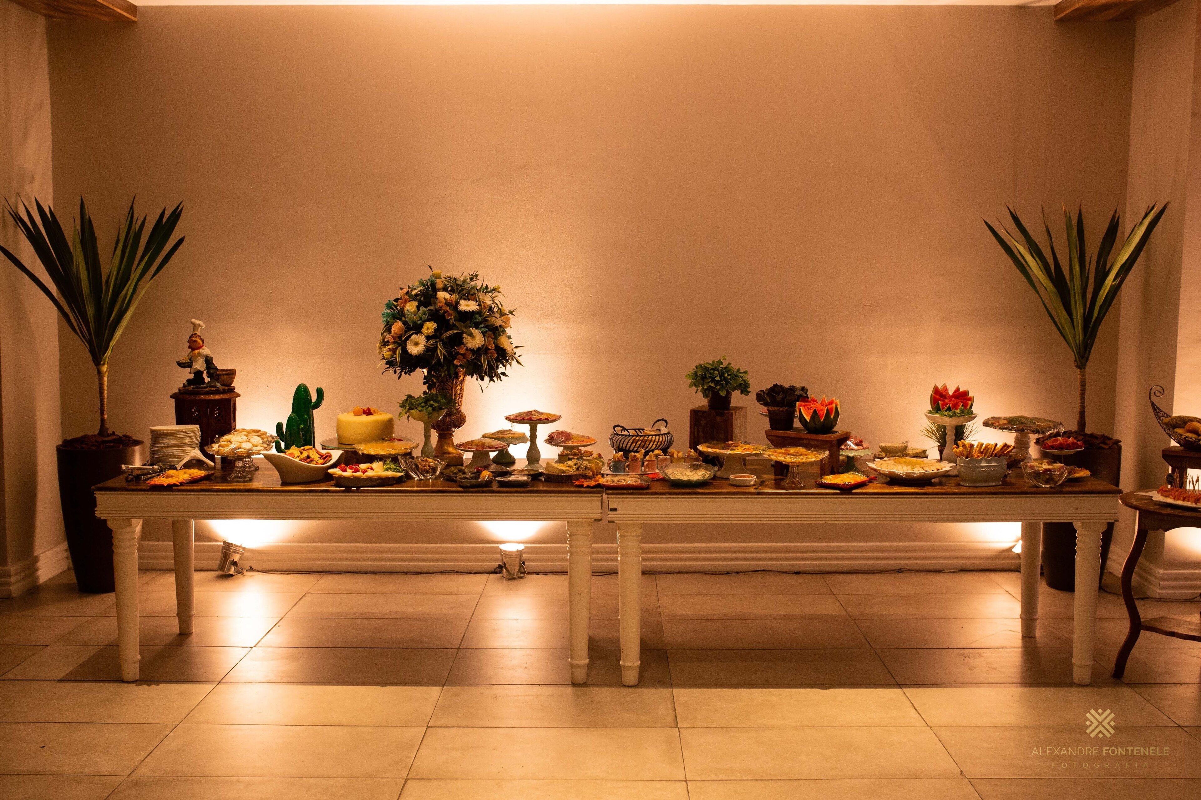 A long dining table with various dishes and centerpieces, flanked by potted plants.