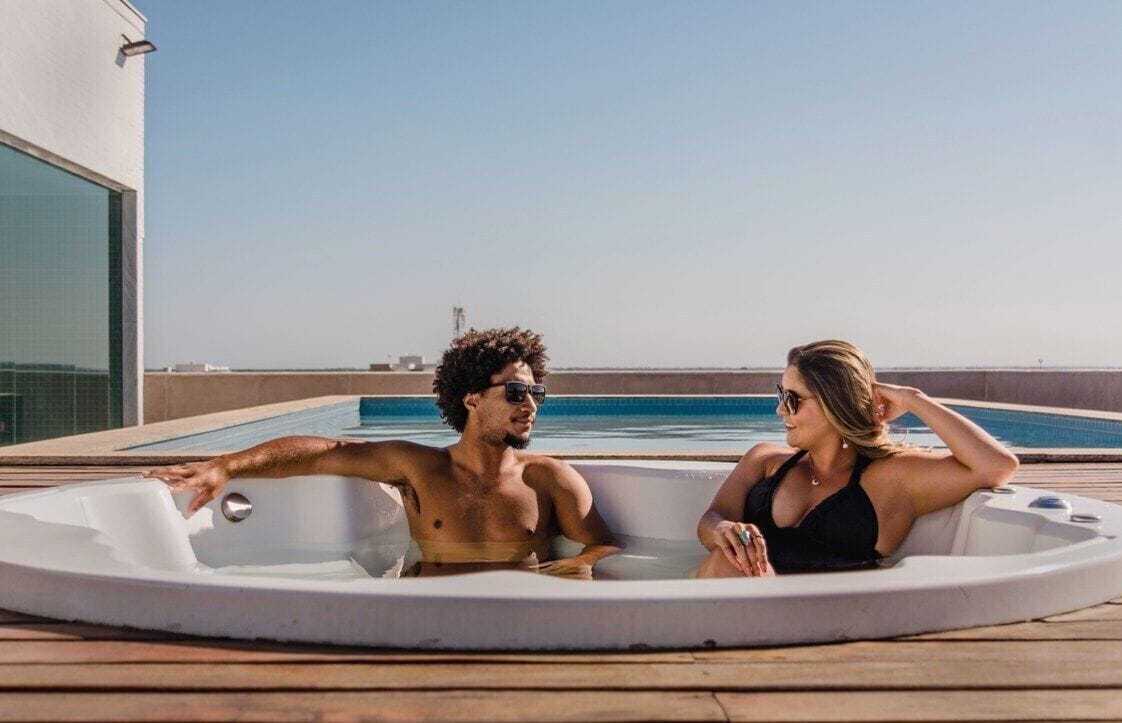Una pareja relajándose en un jacuzzi sobre una terraza de madera, con vista despejada al mar y un edificio al fondo.