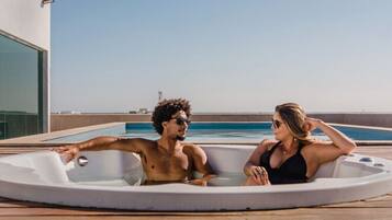 Una pareja relajándose en un jacuzzi sobre una terraza de madera, con vista despejada al mar y un edificio al fondo.