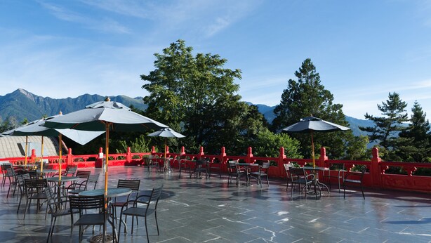 En udendørs terrasse med borde, stole, parasoller og et rødt gelænder.