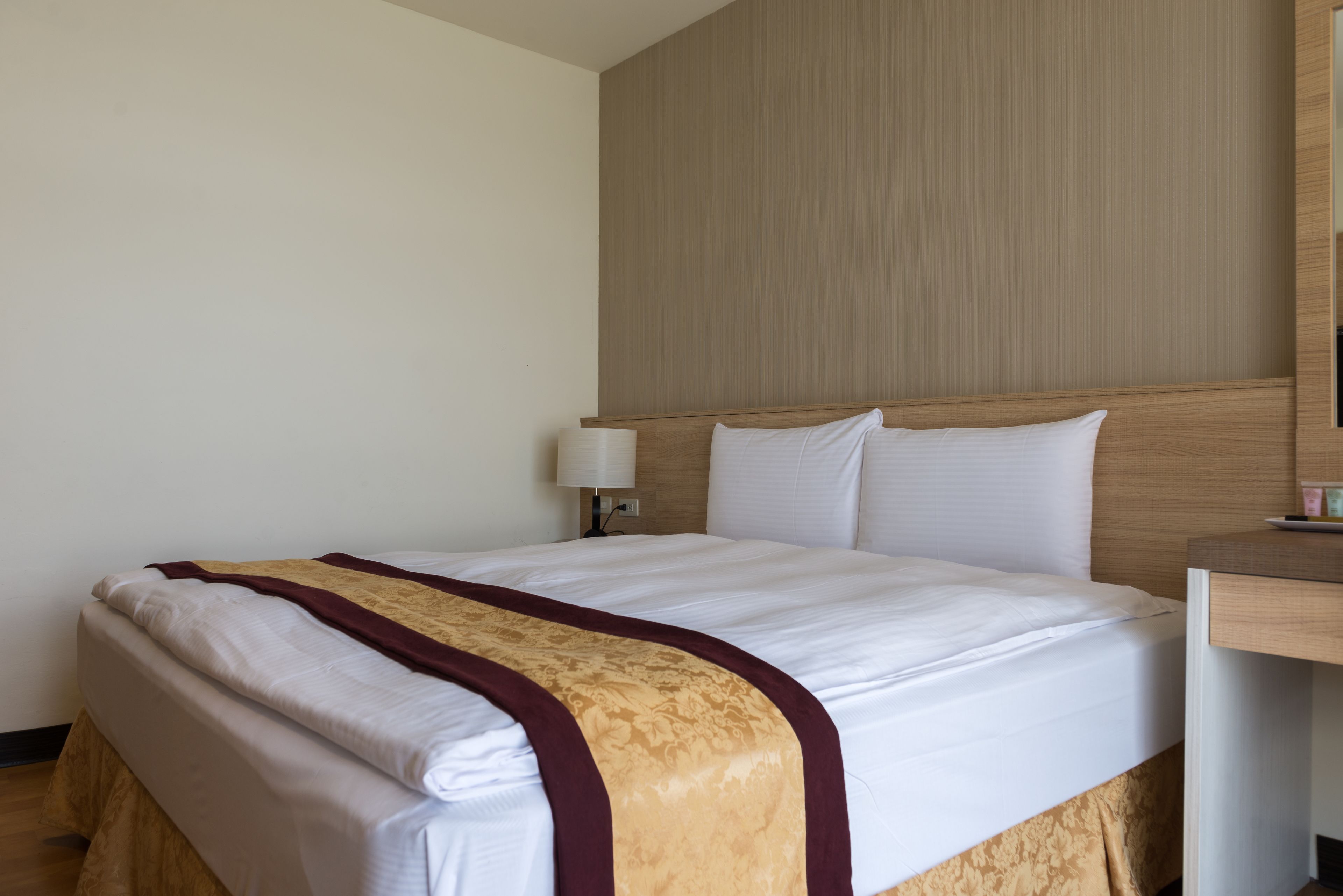 A hotel room with a neatly made bed, a bedside table with a lamp, and a wooden headboard.