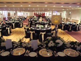 Un salón de banquetes con mesas redondas, manteles negros y centros de mesa con flores blancas.