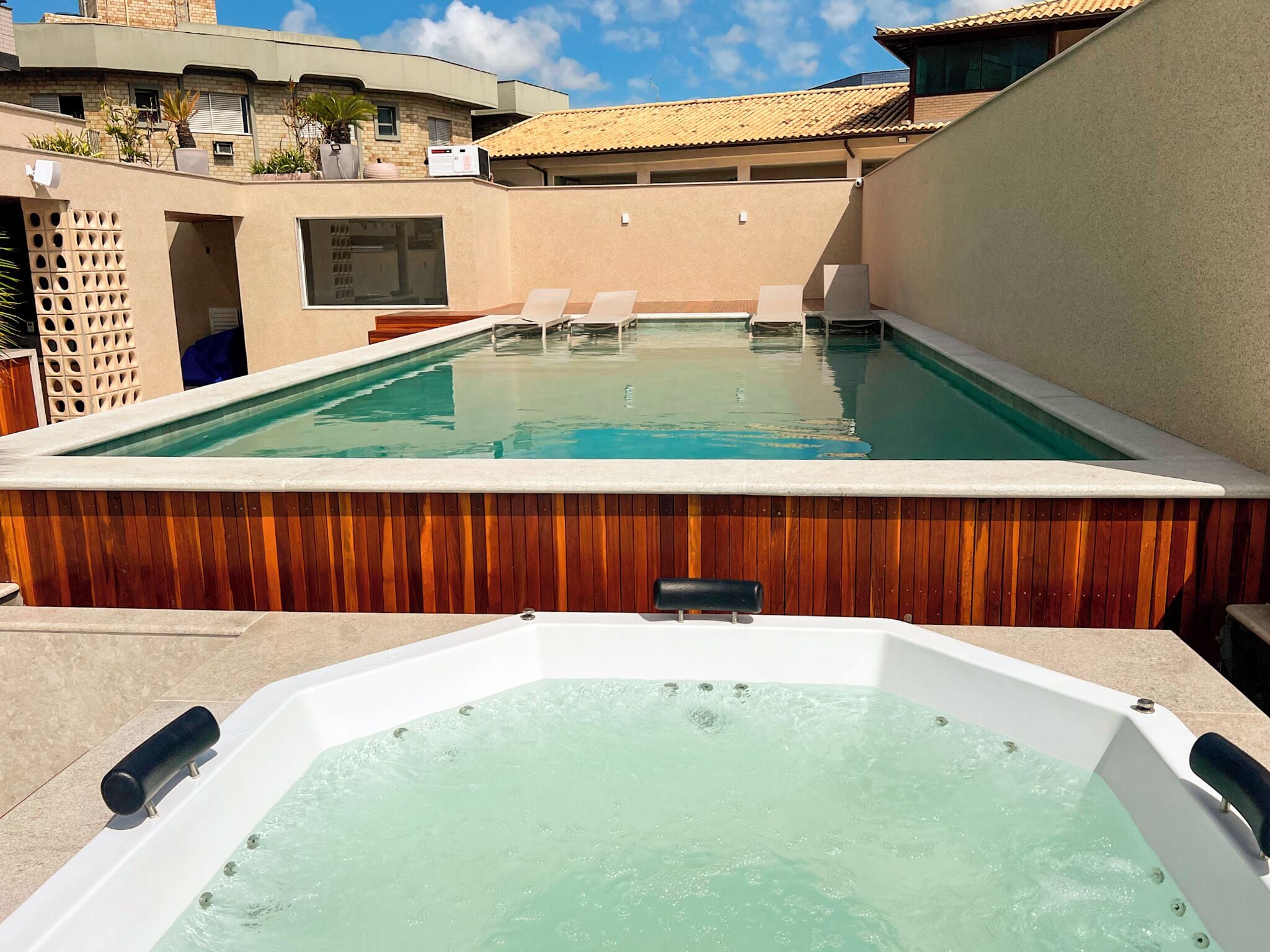 Uma banheira de hidromassagem com estrutura de madeira, piscina e um edifício com telhado de azulejos ao fundo.
