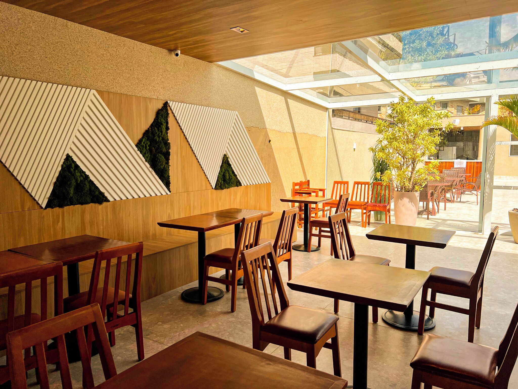 A modern indoor dining area with wooden tables and chairs, a geometric wall design, and a glass ceiling allowing natural light.