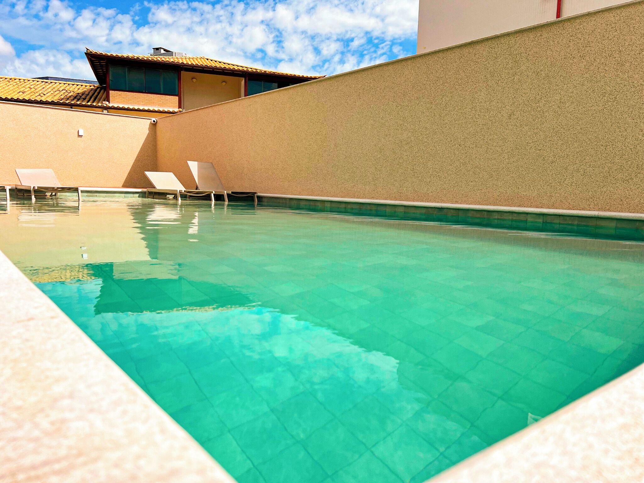 A swimming pool with clear water, surrounded by beige walls and a building with a tiled roof.