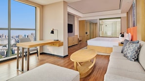 A modern hotel room with a large window, a sofa, a dining table, a bed, and a bathroom with a glass shower. - Hilton Garden Inn Wuhan Hankou (Wuhan)