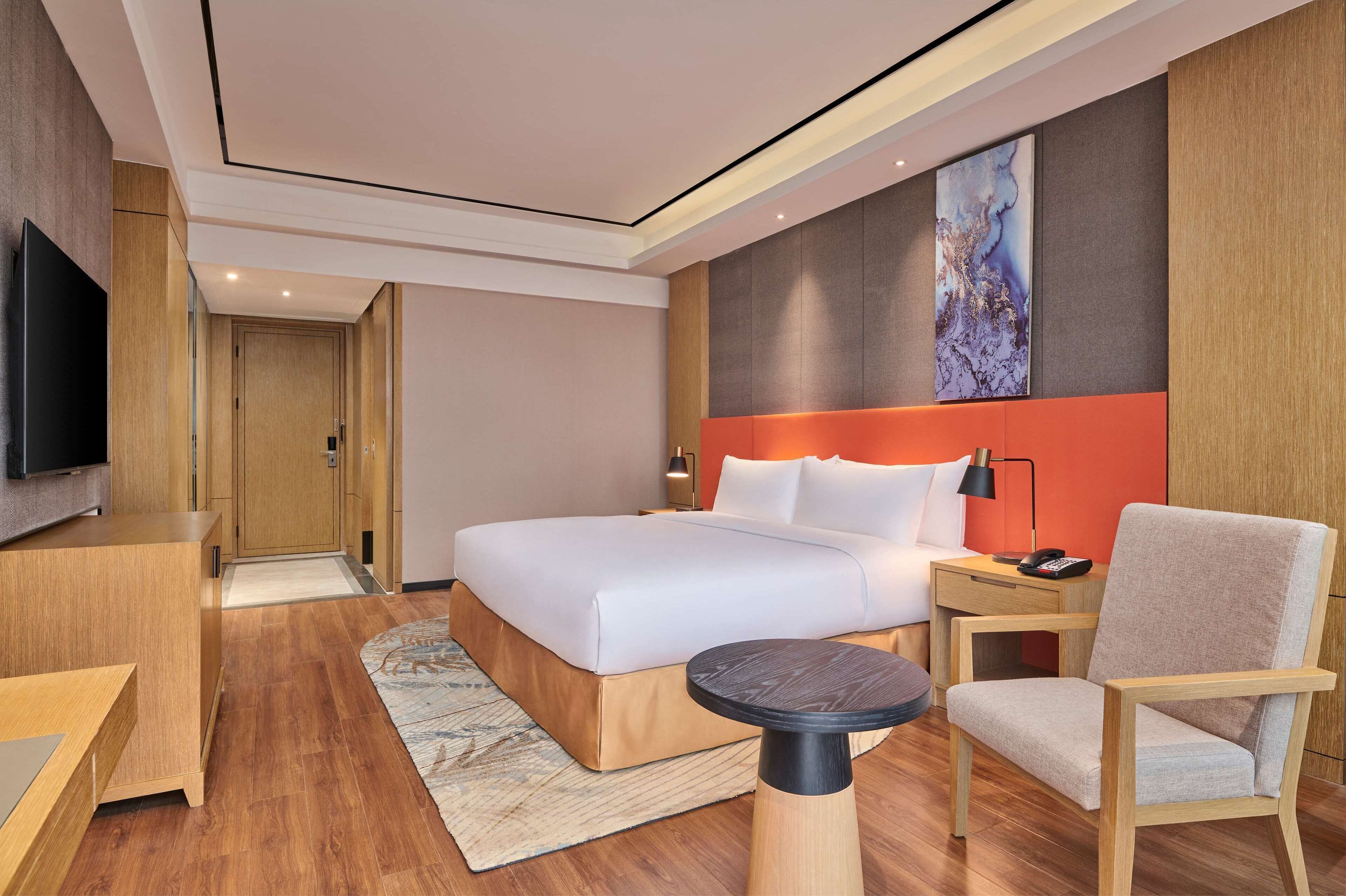 A modern hotel room with a large bed, a desk, a chair, a TV, and a painting on the wall.