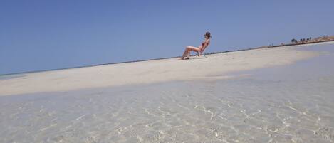 Una playa con agua clara y poco profunda, y una persona sentada en una silla de playa.