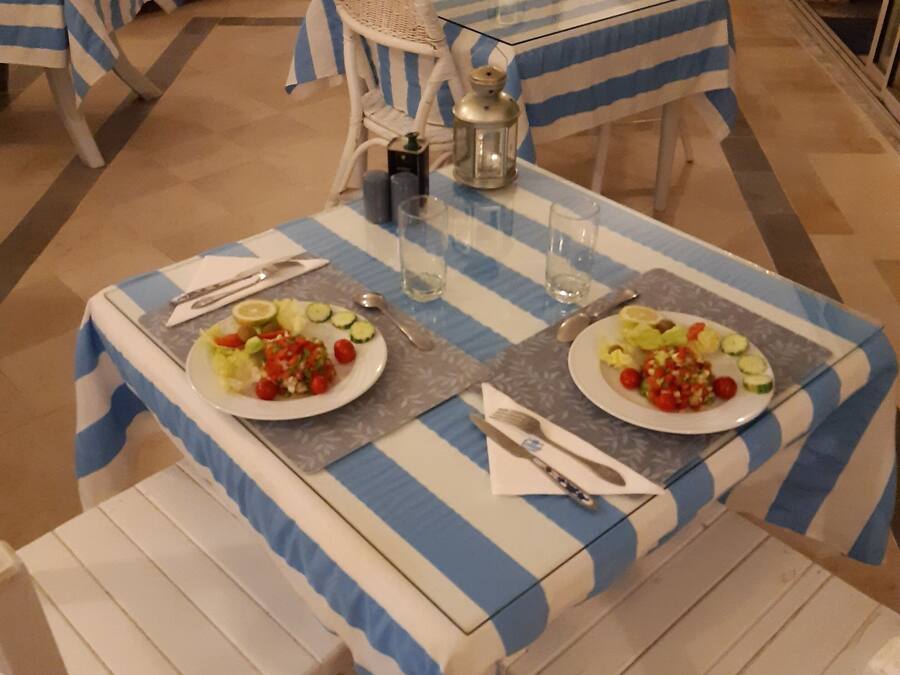 A dining area with two tables set for two, each with a blue and white striped tablecloth, white wicker chairs, and plates of salad.