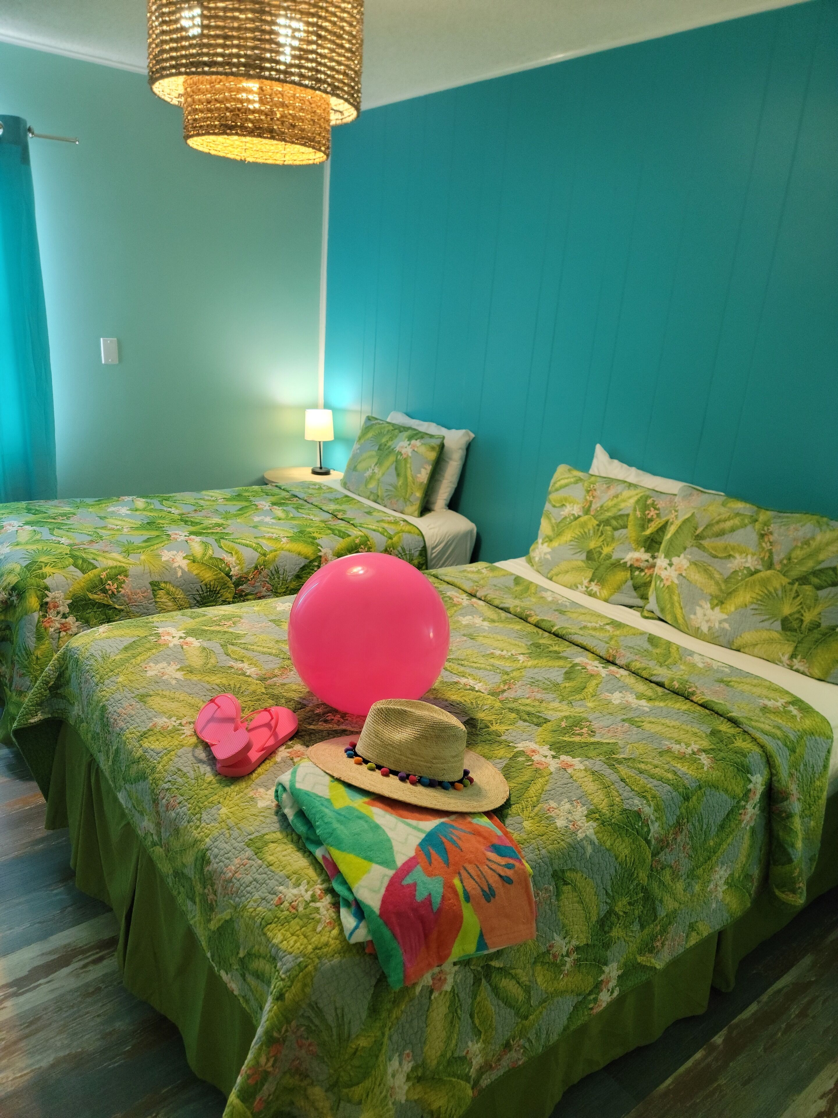 Een kamer met blauwgroene wanden, twee bedden met bloemenbedrukking, een roze ballon, een strooien hoed en een kleurrijke handdoek.