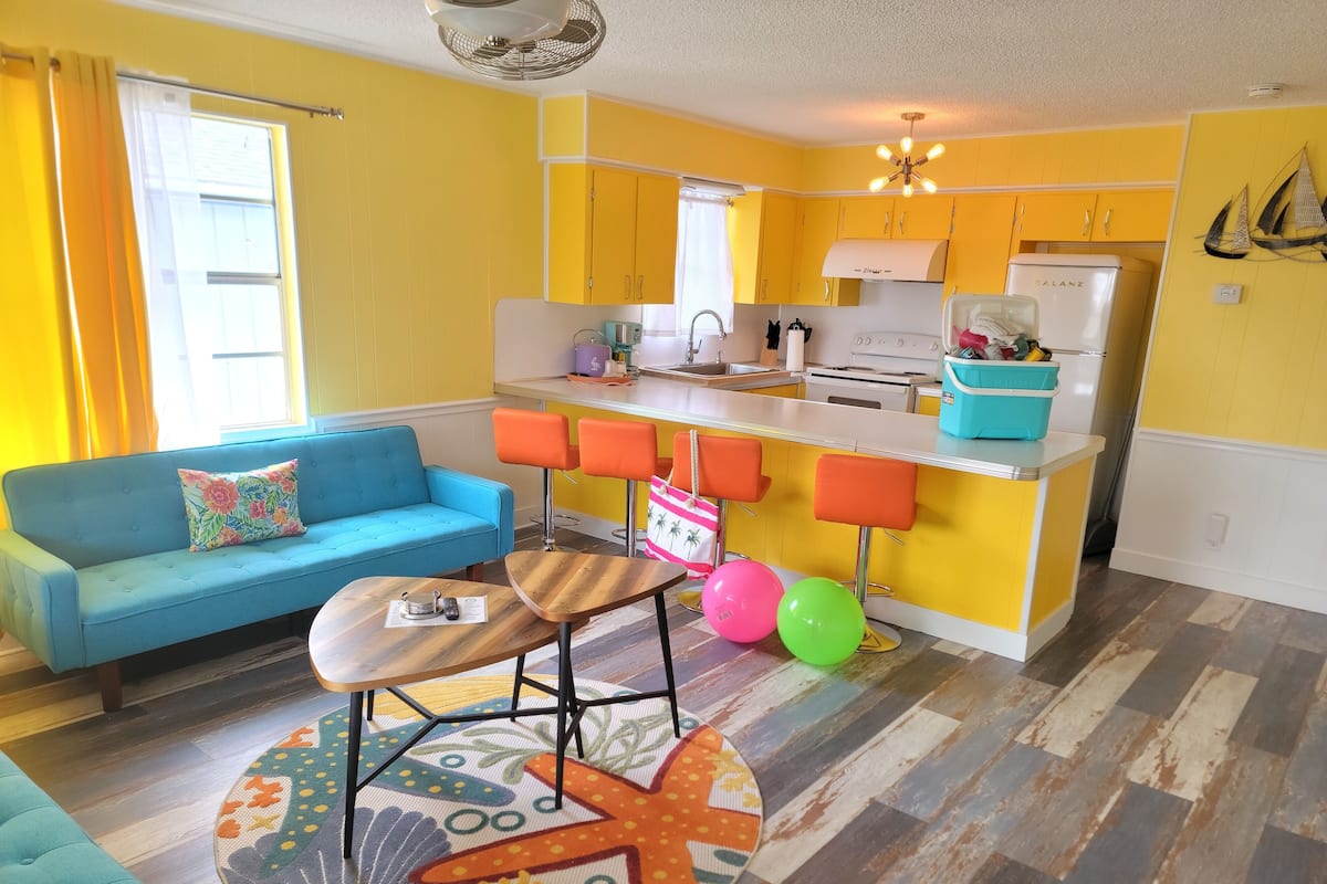 A compact living space with a blue sofa, a small round table, and a kitchen area featuring yellow cabinets and a white refrigerator.